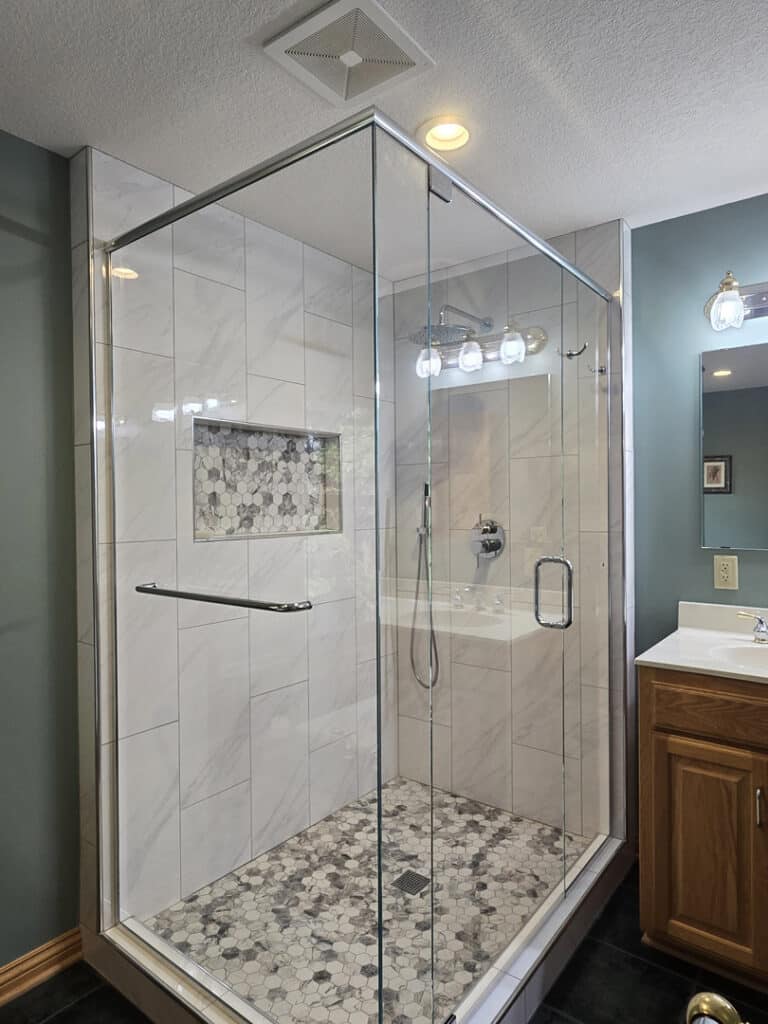 Frameless glass shower enclosure with chrome hardware and marble tile walls and hex tile floor