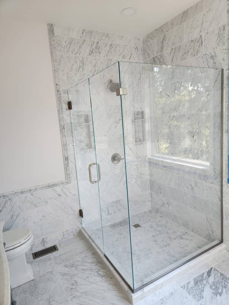 Large frameless glass shower enclosure with marble tile, window view, and minimalist design